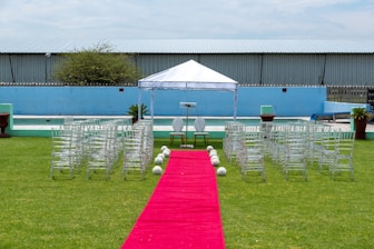 A wedding ceremony setup featuring a white canopy tent at the center with transparent chairs arranged in rows on a green lawn. A red carpet aisle is flanked by white flower arrangements, leading to the canopy. The background includes a blue painted wall, some industrial buildings, and potted plants.