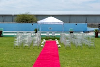 A wedding ceremony setup featuring a white canopy tent at the center with transparent chairs arranged in rows on a green lawn. A red carpet aisle is flanked by white flower arrangements, leading to the canopy. The background includes a blue painted wall, some industrial buildings, and potted plants.