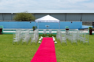 A wedding ceremony setup featuring a white canopy tent at the center with transparent chairs arranged in rows on a green lawn. A red carpet aisle is flanked by white flower arrangements, leading to the canopy. The background includes a blue painted wall, some industrial buildings, and potted plants.