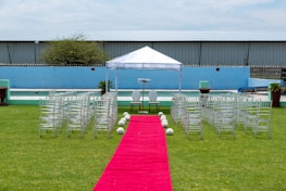 A wedding ceremony setup featuring a white canopy tent at the center with transparent chairs arranged in rows on a green lawn. A red carpet aisle is flanked by white flower arrangements, leading to the canopy. The background includes a blue painted wall, some industrial buildings, and potted plants.