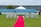 A wedding ceremony setup featuring a white canopy tent at the center with transparent chairs arranged in rows on a green lawn. A red carpet aisle is flanked by white flower arrangements, leading to the canopy. The background includes a blue painted wall, some industrial buildings, and potted plants.