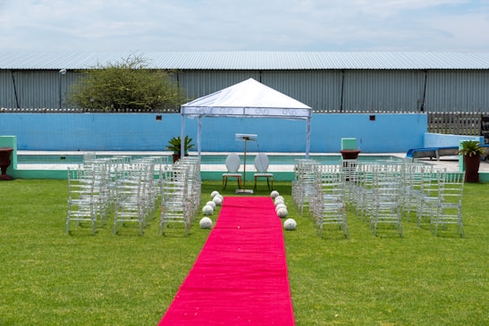 A wedding ceremony setup featuring a white canopy tent at the center with transparent chairs arranged in rows on a green lawn. A red carpet aisle is flanked by white flower arrangements, leading to the canopy. The background includes a blue painted wall, some industrial buildings, and potted plants.