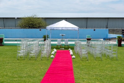 A wedding ceremony setup featuring a white canopy tent at the center with transparent chairs arranged in rows on a green lawn. A red carpet aisle is flanked by white flower arrangements, leading to the canopy. The background includes a blue painted wall, some industrial buildings, and potted plants.