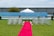 A wedding ceremony setup featuring a white canopy tent at the center with transparent chairs arranged in rows on a green lawn. A red carpet aisle is flanked by white flower arrangements, leading to the canopy. The background includes a blue painted wall, some industrial buildings, and potted plants.