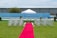 A wedding ceremony setup featuring a white canopy tent at the center with transparent chairs arranged in rows on a green lawn. A red carpet aisle is flanked by white flower arrangements, leading to the canopy. The background includes a blue painted wall, some industrial buildings, and potted plants.