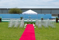 A wedding ceremony setup featuring a white canopy tent at the center with transparent chairs arranged in rows on a green lawn. A red carpet aisle is flanked by white flower arrangements, leading to the canopy. The background includes a blue painted wall, some industrial buildings, and potted plants.