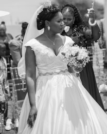 A bride in a decorative white wedding dress holding a bouquet of flowers stands outdoors, surrounded by people attending the ceremony. Her hair is styled elegantly, and she wears a veil and a tiara.