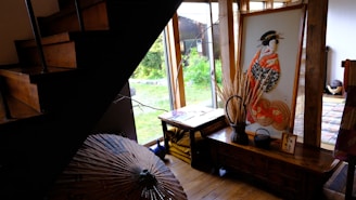 A cozy interior space featuring a wooden staircase and a corner decorated with a large framed artwork depicting a traditional Japanese figure in a kimono. The room has a wooden floor and is well-lit by natural light from large windows. Various decorative items like a parasol, a small table, and a woven basket with dried plants are present, adding an artistic and cultural ambiance.