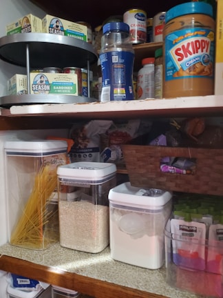 A pantry with organized items including jars of peanut butter, canned sardines, dry pasta, rice, sugar, and various packaged goods. A spice carousel with boxed and canned foods is also visible.