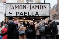 A busy street market scene with a food stall selling paella. The stall has a sign reading 'Jamon Jamon PAELLA'. A crowd of people, dressed in winter clothing, gathers around the stall. The atmosphere is lively as customers engage with the sellers.
