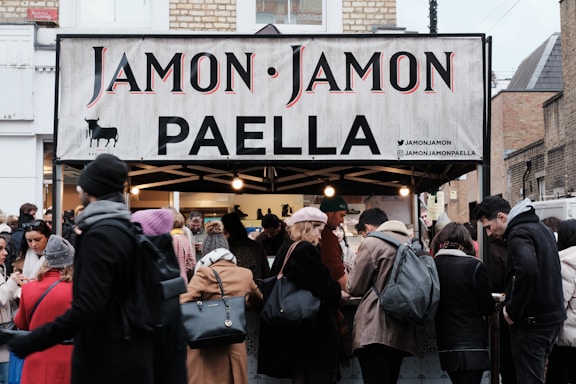 A busy street market scene with a food stall selling paella. The stall has a sign reading 'Jamon Jamon PAELLA'. A crowd of people, dressed in winter clothing, gathers around the stall. The atmosphere is lively as customers engage with the sellers.