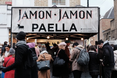 A busy street market scene with a food stall selling paella. The stall has a sign reading 'Jamon Jamon PAELLA'. A crowd of people, dressed in winter clothing, gathers around the stall. The atmosphere is lively as customers engage with the sellers.