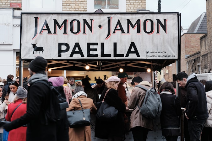 A busy street market scene with a food stall selling paella. The stall has a sign reading 'Jamon Jamon PAELLA'. A crowd of people, dressed in winter clothing, gathers around the stall. The atmosphere is lively as customers engage with the sellers.