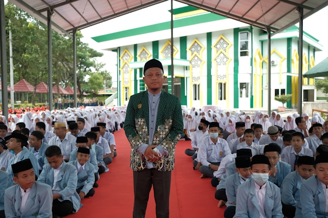 A large gathering of students sitting on a red carpet, with a man standing prominently in front, wearing a green patterned jacket and a black cap. The background features a colorful building with green, yellow, and white accents, resembling a mosque or religious school. Many of the students are dressed in light blue blazers and white shirts, and some are wearing headscarves, indicating a formal or religious setting.