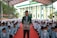 A large gathering of students sitting on a red carpet, with a man standing prominently in front, wearing a green patterned jacket and a black cap. The background features a colorful building with green, yellow, and white accents, resembling a mosque or religious school. Many of the students are dressed in light blue blazers and white shirts, and some are wearing headscarves, indicating a formal or religious setting.