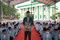 A large gathering of students sitting on a red carpet, with a man standing prominently in front, wearing a green patterned jacket and a black cap. The background features a colorful building with green, yellow, and white accents, resembling a mosque or religious school. Many of the students are dressed in light blue blazers and white shirts, and some are wearing headscarves, indicating a formal or religious setting.