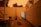 A backyard with an open door leading to an indoor space. There are several bicycles scattered around, a yoga mat is on the ground, and a potted plant with wilting leaves. The lighting is dim with an orange hue, likely from an evening or fire-related sky.