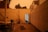 A backyard with an open door leading to an indoor space. There are several bicycles scattered around, a yoga mat is on the ground, and a potted plant with wilting leaves. The lighting is dim with an orange hue, likely from an evening or fire-related sky.