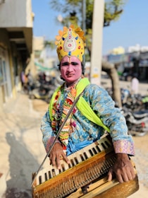 A person dressed in traditional, vibrant attire, including a decorated headdress and colorful garments, playing a harmonium outdoors with a street scene in the background.