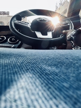 The image captures the interior of a car, viewed from a low angle. The focus is on the steering wheel, central dashboard, and a hand gripping the steering wheel. The sun is shining through the windshield, creating a glare effect on the steering wheel. The bottom part of the image shows a denim-clad leg, adding a casual feel to the scene.