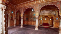 A richly decorated ornate room featuring intricately carved pillars and colorful archways. The space is adorned with detailed floral patterns and motifs in vibrant reds, golds, and browns. A plush seating area is tucked under an ornate arch, reflecting a luxurious and historical architectural style.