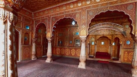 A richly decorated ornate room featuring intricately carved pillars and colorful archways. The space is adorned with detailed floral patterns and motifs in vibrant reds, golds, and browns. A plush seating area is tucked under an ornate arch, reflecting a luxurious and historical architectural style.