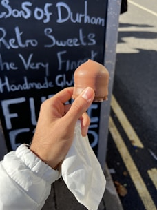 A hand holding a chocolate-covered treat is depicted, with a blackboard in the background displaying handwritten text about sweets and gluten-free products. The person is wearing a white jacket, and the scene takes place on a sunny day, with part of a road visible in the background.