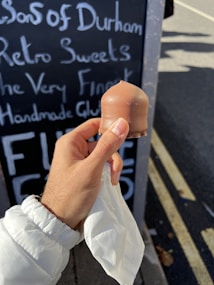 A hand holding a chocolate-covered treat is depicted, with a blackboard in the background displaying handwritten text about sweets and gluten-free products. The person is wearing a white jacket, and the scene takes place on a sunny day, with part of a road visible in the background.