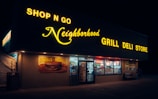A convenience store illuminated by bright yellow and white neon signage at night, featuring a sign reading 'Shop N Go Neighborhood Grill Deli Store'. The front of the store displays a poster advertising 'Homemade Freshness' with a picture of a burger, and various items and promotions visible through the windows.
