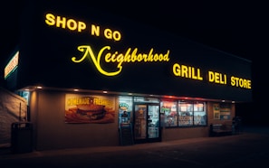 A convenience store illuminated by bright yellow and white neon signage at night, featuring a sign reading 'Shop N Go Neighborhood Grill Deli Store'. The front of the store displays a poster advertising 'Homemade Freshness' with a picture of a burger, and various items and promotions visible through the windows.