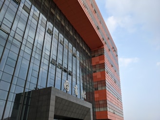 A modern building with a prominent facade composed of glass panels reflecting the sky, interspersed with bold, geometric sections made of red-orange material. The architecture features clean, vertical lines and a protruding entrance with signage in a stylized script.