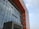 A modern building with a prominent facade composed of glass panels reflecting the sky, interspersed with bold, geometric sections made of red-orange material. The architecture features clean, vertical lines and a protruding entrance with signage in a stylized script.