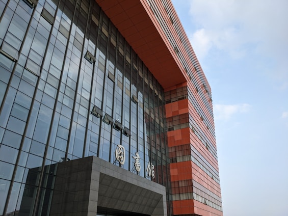 A modern building with a prominent facade composed of glass panels reflecting the sky, interspersed with bold, geometric sections made of red-orange material. The architecture features clean, vertical lines and a protruding entrance with signage in a stylized script.