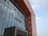A modern building with a prominent facade composed of glass panels reflecting the sky, interspersed with bold, geometric sections made of red-orange material. The architecture features clean, vertical lines and a protruding entrance with signage in a stylized script.