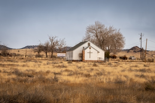 A small white chapel with a large cross on its facade is situated in an open field of dry grass. Bare trees surround the building, and distant mountains are visible under a cloudy sky. A sign near the building and several power lines and poles are present, contributing to a rural setting.