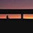A silhouette of a person with a ponytail is visible against a vibrant sunset, seen through a horizontal window frame. The sky transitions from deep purple at the top to orange and yellow near the horizon.