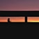 A silhouette of a person with a ponytail is visible against a vibrant sunset, seen through a horizontal window frame. The sky transitions from deep purple at the top to orange and yellow near the horizon.