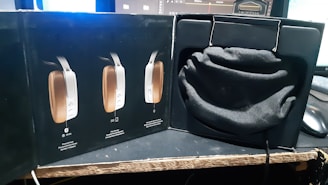 A product box is opened to reveal a soft black pouch resting inside a compartment. The outer section of the box displays images of a sleek, modern electronic device with controls on the side. The setup is on a table with computer monitors in the background.
