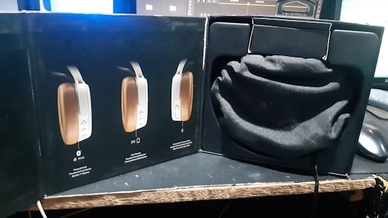 A product box is opened to reveal a soft black pouch resting inside a compartment. The outer section of the box displays images of a sleek, modern electronic device with controls on the side. The setup is on a table with computer monitors in the background.