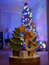 A handcrafted wooden figure of an angel holding a star stands next to a small, decorated evergreen tree. In front of the angel is a gift box with a polka dot pattern. In the background, a beautifully lit and decorated Christmas tree with colorful lights adds a festive atmosphere.