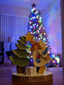 A handcrafted wooden figure of an angel holding a star stands next to a small, decorated evergreen tree. In front of the angel is a gift box with a polka dot pattern. In the background, a beautifully lit and decorated Christmas tree with colorful lights adds a festive atmosphere.