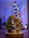 A handcrafted wooden figure of an angel holding a star stands next to a small, decorated evergreen tree. In front of the angel is a gift box with a polka dot pattern. In the background, a beautifully lit and decorated Christmas tree with colorful lights adds a festive atmosphere.