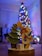 A handcrafted wooden figure of an angel holding a star stands next to a small, decorated evergreen tree. In front of the angel is a gift box with a polka dot pattern. In the background, a beautifully lit and decorated Christmas tree with colorful lights adds a festive atmosphere.
