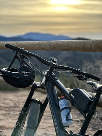 A mountain bike is positioned in front of a wooden fence with a scenic mountainous landscape in the background. A bicycle helmet is hanging from the handlebars, and a water bottle is securely placed on the bike frame. The backdrop features a serene sunset with a blend of warm and cool colors across the sky.