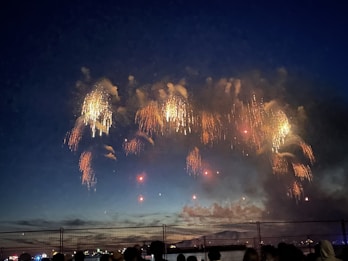 Bright, colorful fireworks explode against a deep blue evening sky. The fireworks create cascading trails of light in various shades of orange and yellow. Below, silhouettes of people observe the display from behind a fence, with a hint of city lights visible in the background.