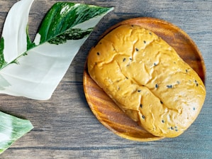 A loaf of bread with black sesame seeds rests on a wooden plate, next to large green and white variegated leaves on a textured wooden surface.