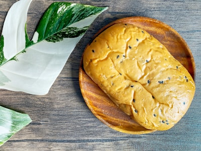 A loaf of bread with black sesame seeds rests on a wooden plate, next to large green and white variegated leaves on a textured wooden surface.