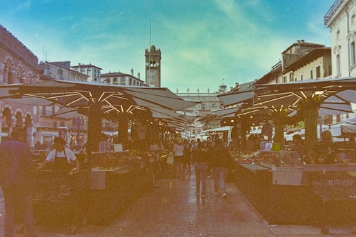 A bustling outdoor market scene with stalls on either side covered by geometric canopies. People are walking through the market, which is set in a historic city square surrounded by Renaissance architecture. A bell tower rises in the background, and the sky above is a bright blue with fluffy clouds.