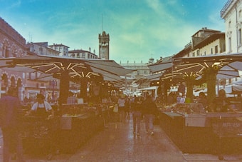 A bustling outdoor market scene with stalls on either side covered by geometric canopies. People are walking through the market, which is set in a historic city square surrounded by Renaissance architecture. A bell tower rises in the background, and the sky above is a bright blue with fluffy clouds.