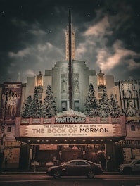 An illuminated theater facade showcasing a marquee sign advertising 'The Book of Mormon' musical. The architecture features art deco elements with intricate designs and ornamentation. The atmosphere is accentuated by dramatic lighting against a cloudy, starry night sky. Christmas trees with lights flank the entrance, adding a festive touch.